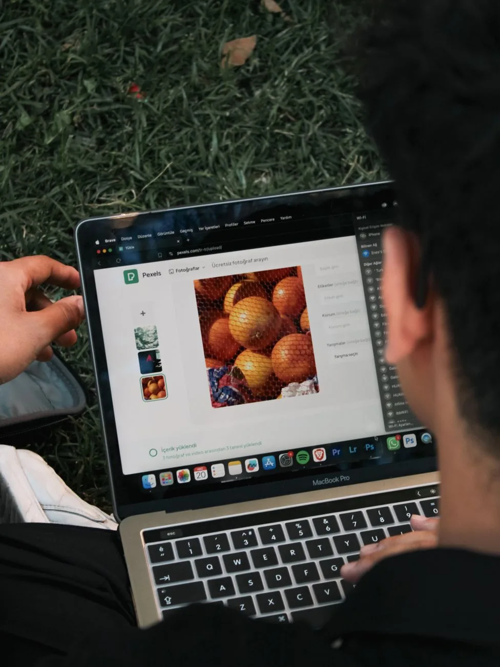 A man edits a fruit image on a MacBook Pro while sitting outdoors on grass.